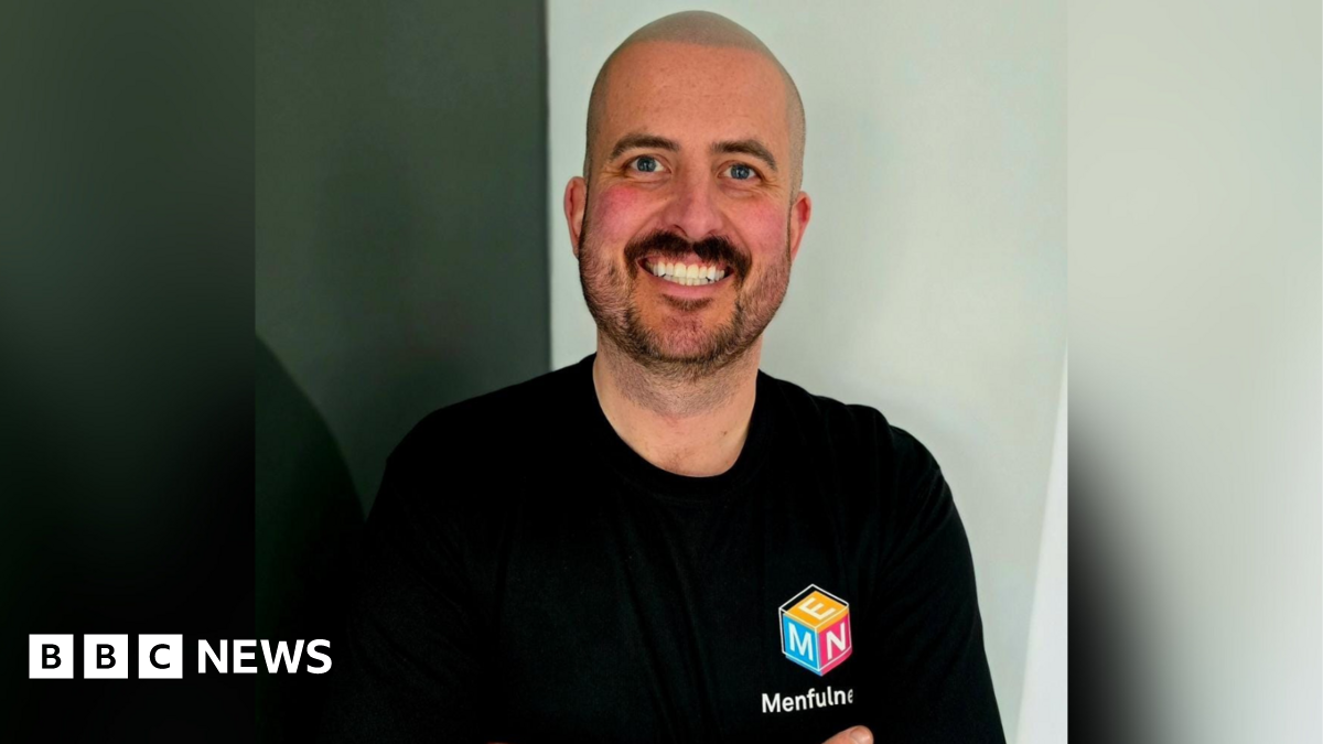 Jack Woodhams smiles at the camera. he has a shaved head and a short, dark beard and wears a black t-shirt with the Menfulness logo on the chest - a cube with blue, orange and pink sides and white lettering below spelling out Menfulness.