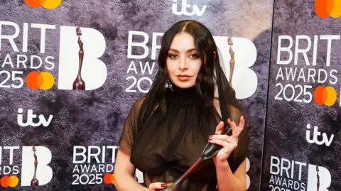PA Charli XCX poses while holding an award in front of a backdrop saying Brit Awards 2025