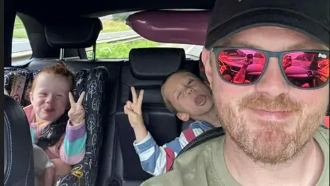 Aled Edwards A man with a ginger beard wearing a black cap, pink tinted sunglasses and a green t shirt. He is sat in a car, and in car seats behind him - both pulling silly faces and making peace signs with their hands - are a young girl with curly ginger hair and a young boy, both wearing striped multi-colour tops.