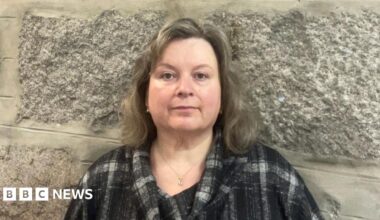 Claire Moggach, a woman with shoulder-length, dark blonde hair wearing a black, grey and white top. She is photographed in front of a granite wall.