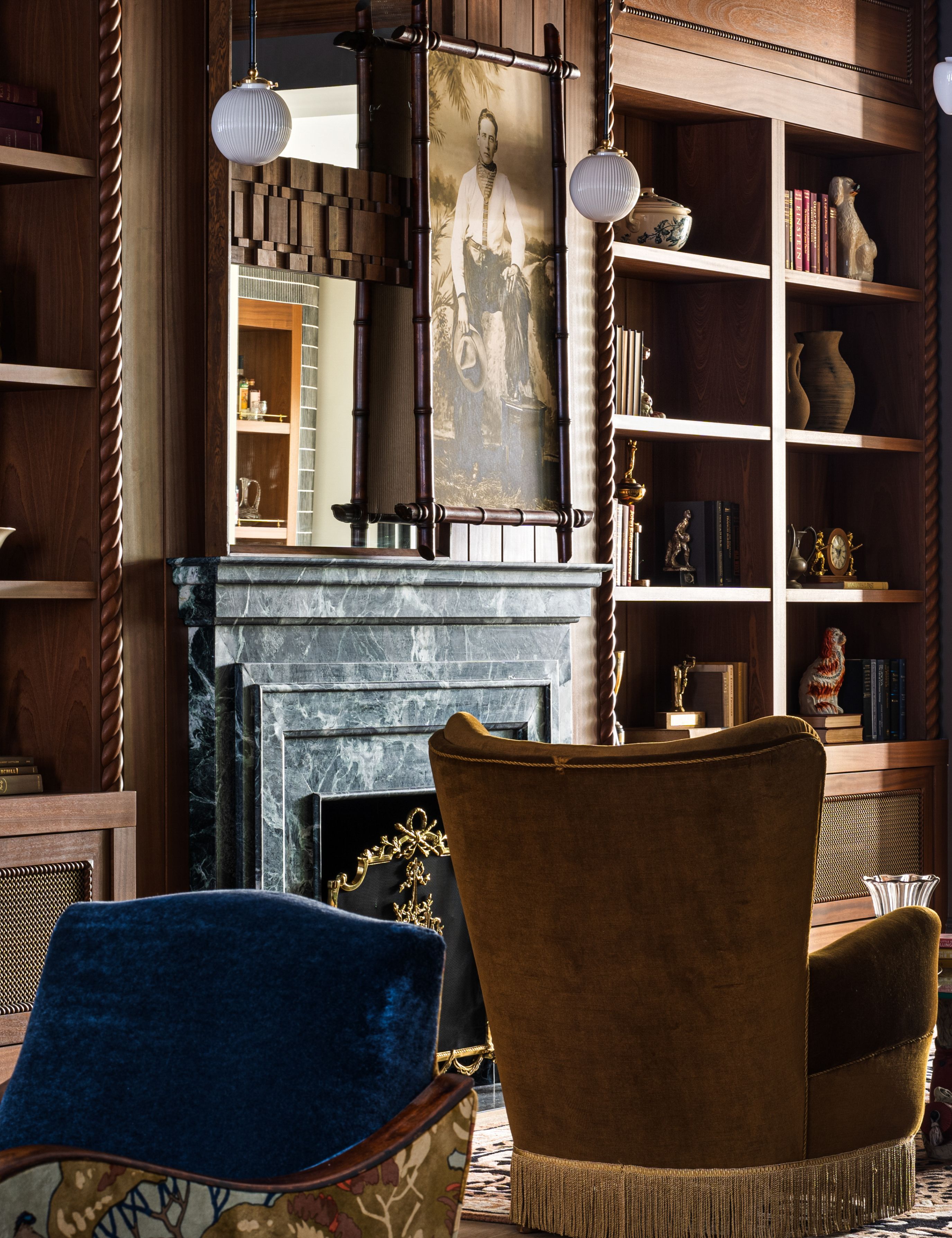 NYC parlor room featuring warm wooden millwork, milk glass pendant lighting, fringed accent chairs, and a striking green marble fireplace