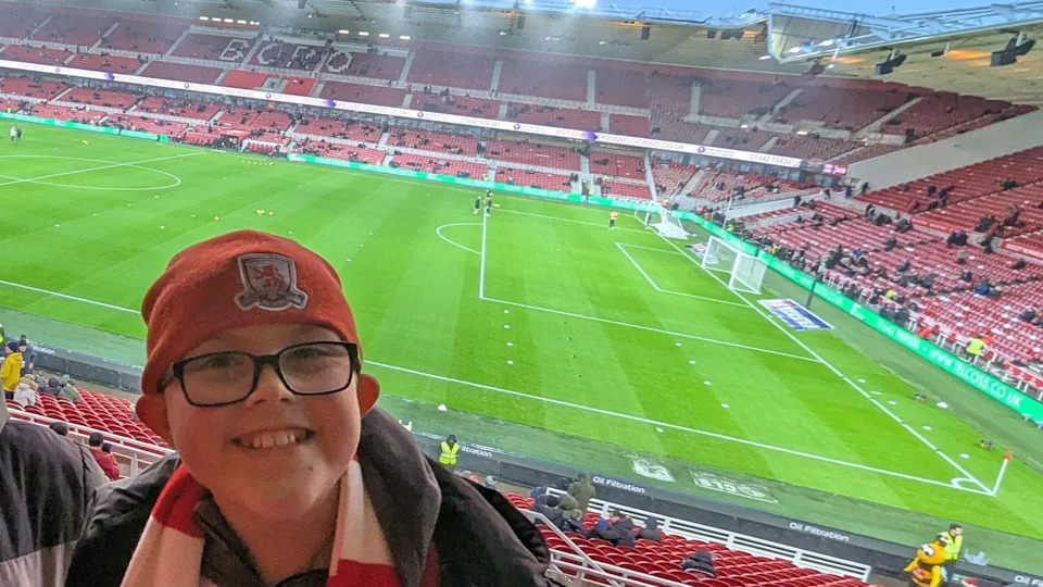 Riley is in a stand at the Riverside Stadium. He is smiling and has his back to the pitch where a few groundstaff are working. The stand on the opposite side and behind one of the goals can be partially seen. There are few fans in as it appears to be well before kick off. Riley is wearing a red Middlesbrough beany hat and red and white striped scarf. He is wearing rectangular black-rimmed glasses and a black coat.
