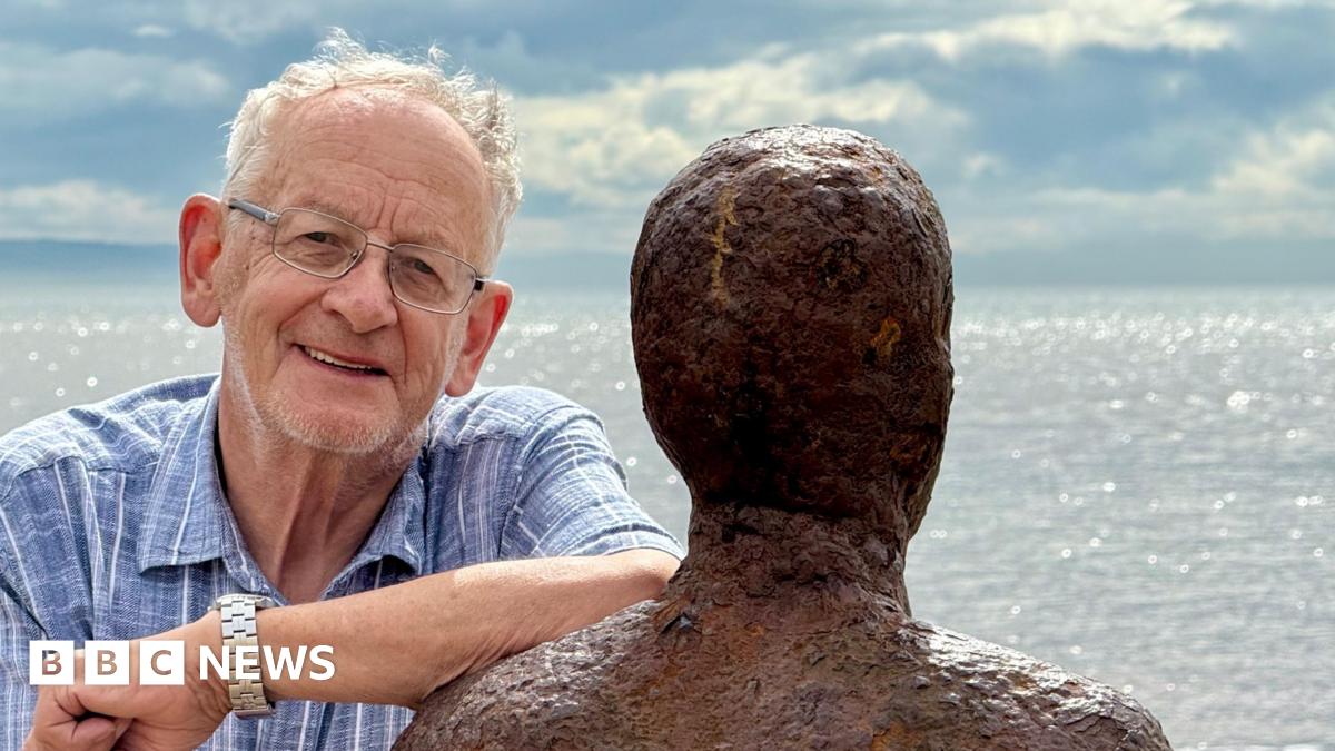 Ron Davies with grey hair wearing a blue and white shirt and glasses leans on one of the Iron Men sculptures on Crosby beach with the incoming tide in the background on a clear day. He is smiling.