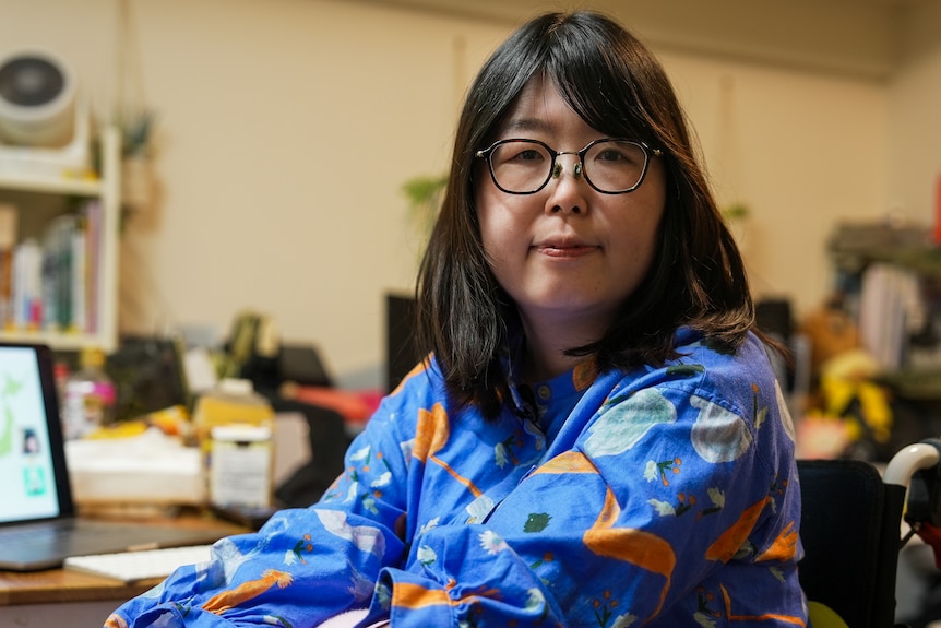 A Japanese woman sits at a desk facing away from her laptop and at the camera. She wears glasses and a blue top.