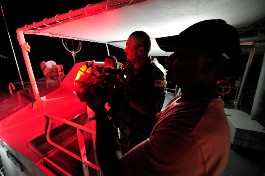 A Navy seaman on a boat instructioning a second seaman holding a large light in his hands