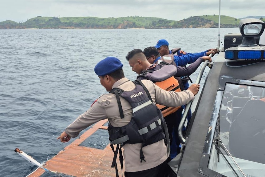 Four men in uniforms and life jackets on the side of a boat looking at a piece of debris floating in water.