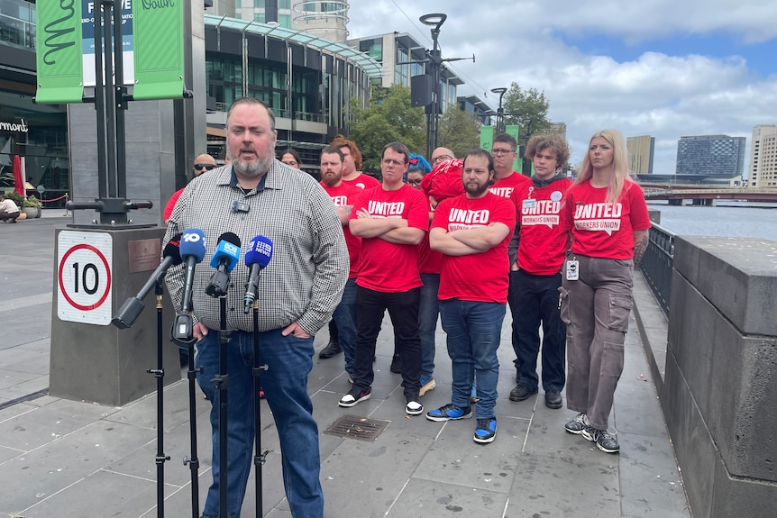 Andrew Jones speaks to media in front of people in red shirts.
