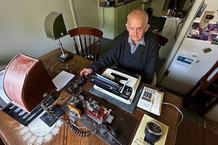 A man sitting at a kitchen table with a typewriter, and Morse code equipment laid out in front of him.