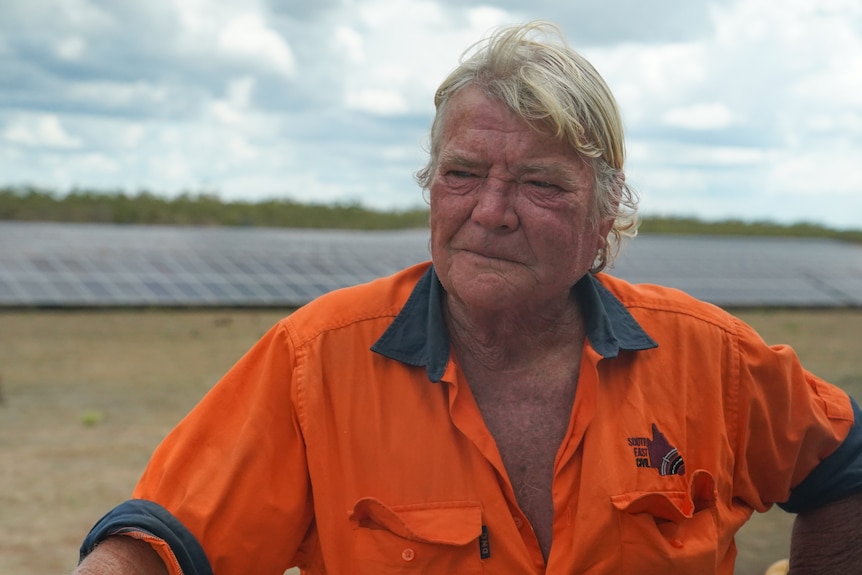 a man with blonde hair wearing a orange hi vis short stands in front of solar panels looking solemn