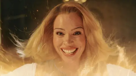 A still shot from the climax of the most recent Doctor Who episode, in which Ncuti Gatwa's Doctor appeared to regenerate into former companion Rose Tyler, played by Billie Piper. The shot shows glimmers and sparks surrounding Rose, as her form is revealed after regenerating. She has a broad, grinning smile with tears forming in her eyes as her blonde hair is flung backwards by the energy surrounding her.