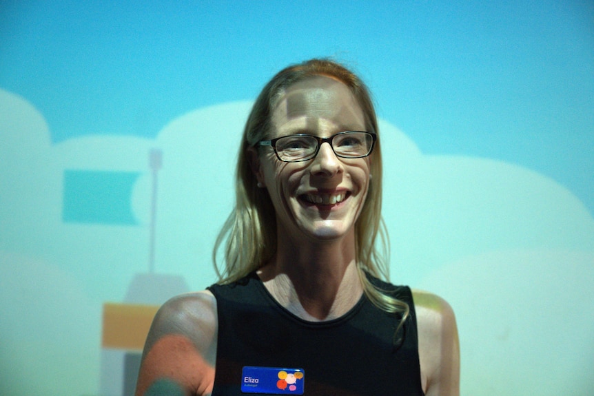 Eliza Brbich, wearing glasses and with long blonde hair, stands smiling against a blue backdrop.