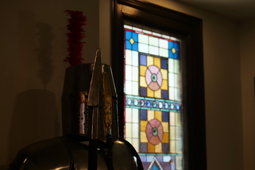 A suit of armour pictured from the chest up, in the background, a colourful lead light window.
