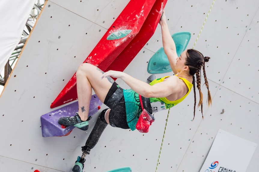 Climber Sarah Larcombe, wearing a gold singlet and braids in her hair, hangs from a red hold on the wall by one hand.