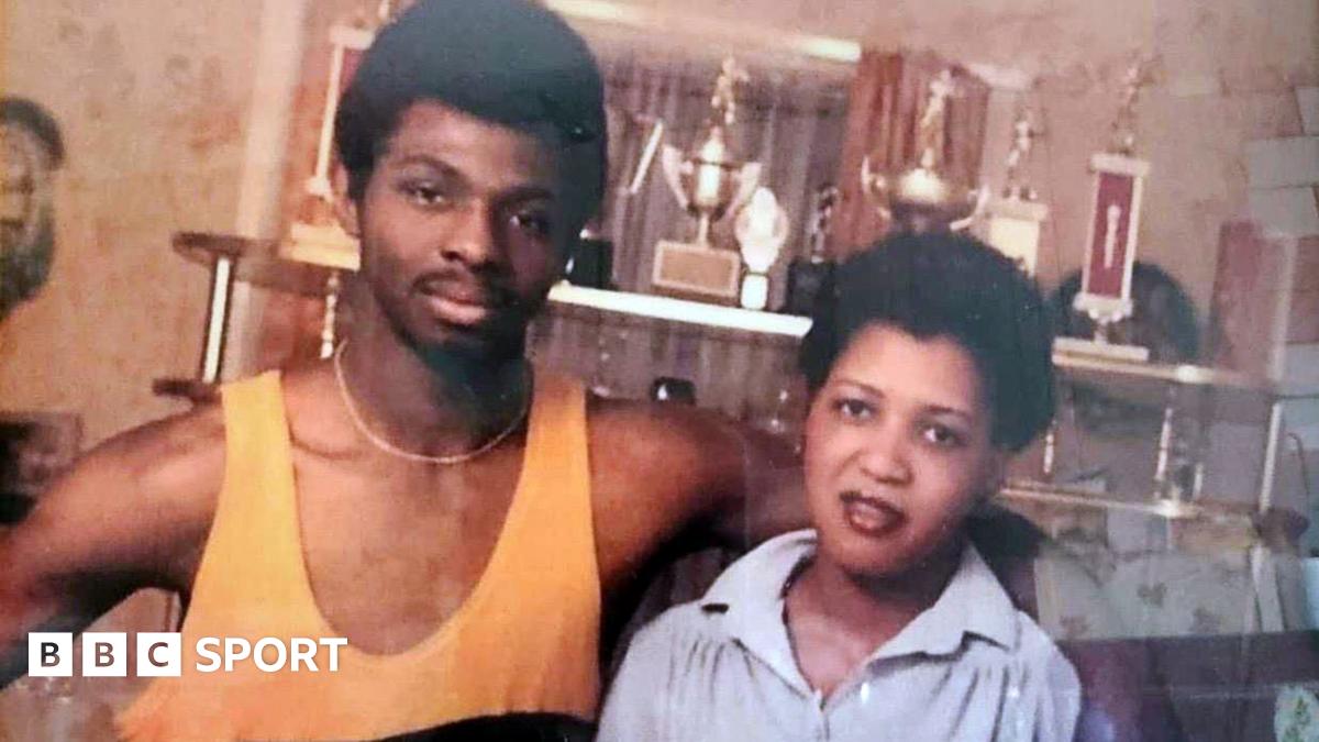 A young James Cook, wearing a yellow boxing vest, with his arm around his wife Carmen. There are various sport trophies behind them.