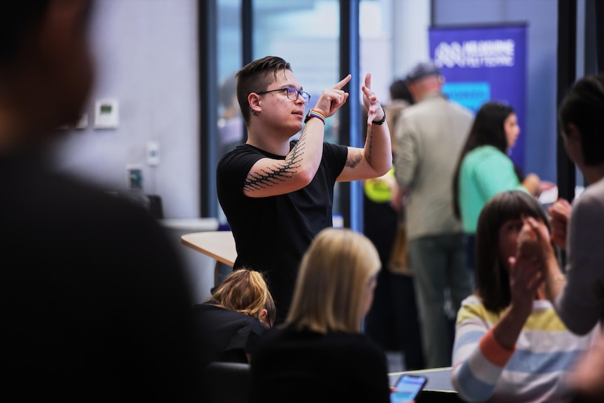 Ryan Malonda, a young man in a black t-shirt uses sign language, gesturing with his hands in the air with people seated nearby.
