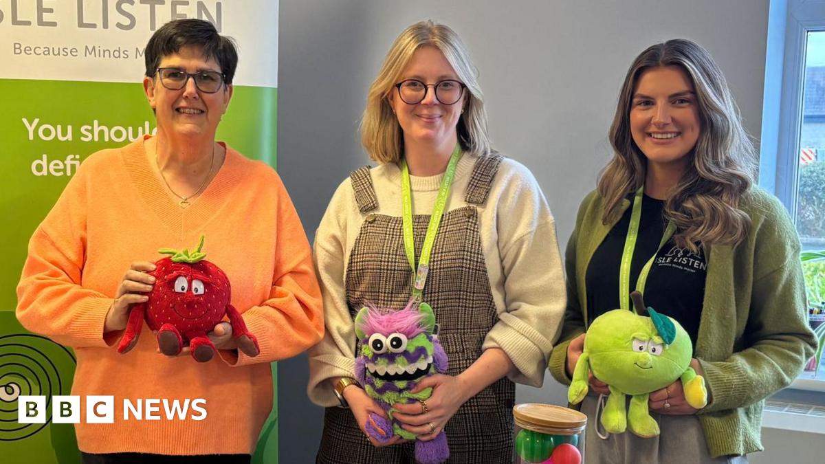 Sarah Kelly from Manx Lottery Trust and Isle Listen's Becks Macnair and Holly Ramsay stand behind the table smiling for the camera. They are holding stuffing children's toys used in the lessons.