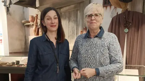Rugby Borough Council A young woman in a blue shirt stood in front of  a medieval display at a museum , next to an older woman with short blonde hair, black glasses and wearing a grey jumper