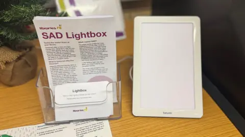 A still image of a Sad lamp sat next to a plastic box containing leaflets about the Sad lightbox. The lamp is an A4-size white light, which is turned off. It is placed on a wooden desk, with the bottom of a small Christmas tree in the distance.