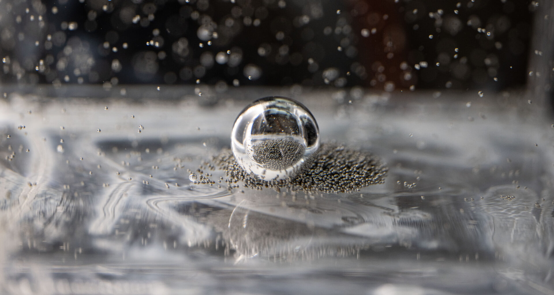 Tiny silver ball bearings crowd around a larger bearing in a clear, viscous fluid. Some of the smaller ball bearings are suspended in the fluid.