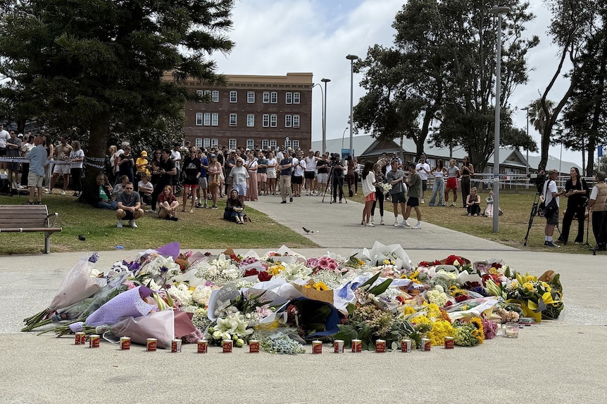 A growing pile of flowers are placed on the ground outside, surrounded by candles