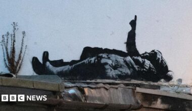 The black and white street art depicts two children lying on their backs and pointing upwards. The child nearest the camera is wearing a wooly hat with hands tucked into their jacket pockets. Behind them a larger person is pointing upwards in a bobbly hat (possibly a Santa type hat) also wearing boots.