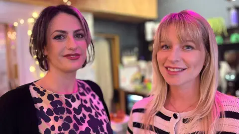 Madelaine Thomas and Jess Davies in a coffee shop smiling at the camera. Madelaine has dark short hair and is wearing an animal print top and black cardigan. Jess has long blonde hair with a  fringe and us wearing a black and white striped cardigan. It is a head and shoulders shot of the two of them.
