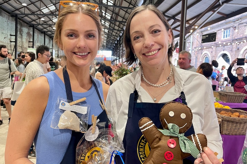 Sonia Martin left in a blue shirt, yellow glasses on her head, smiling with a woman next to her, both at a market holding goods.