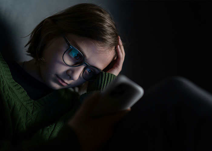 Teenage girl wearing glasses looks distressed while using a smartphone in a dark room, highlighting ChatGPT controversy.