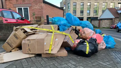 Cardboard boxes and blue, orange and black refuge bags are piled on a street corner. They have yellow environmental crime scene tape stuck across the mess