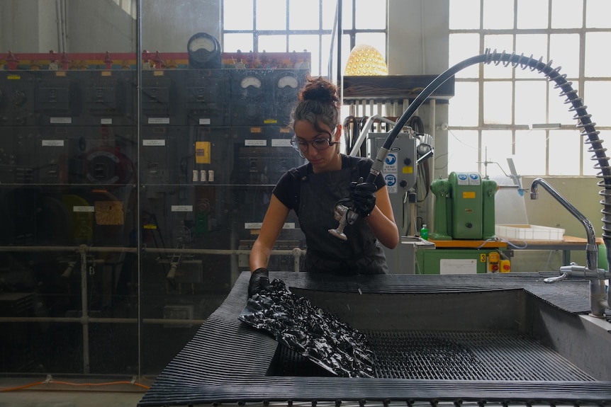 A woman in an apron and gloves hoses down a distorted piece of black glass at an industrial sink.