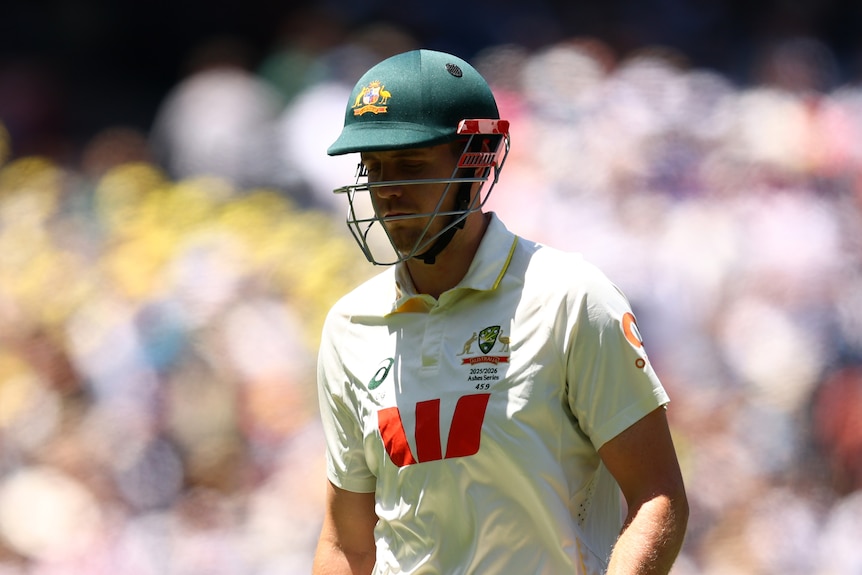 A cricketer wearing a green helmet looks downwards as he walks off the pitch