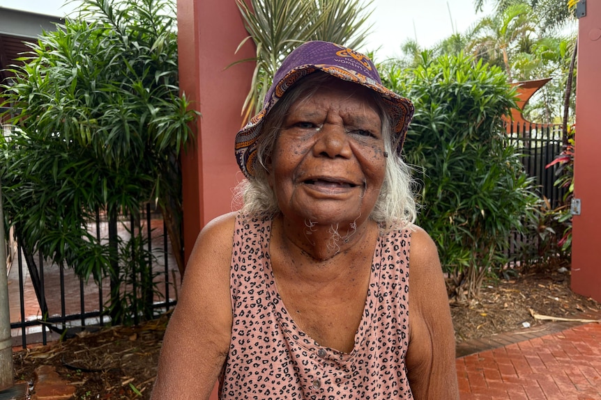 An elderly Aboriginal woman smiles at the camera wearing a hat outdoors.