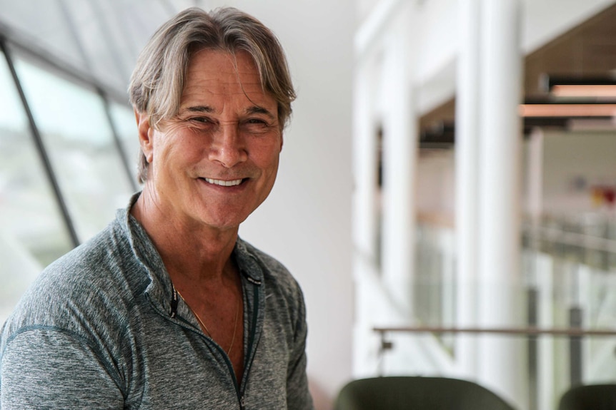 Richard Norton smiling at camera in a grey shirt, in front of a white hallway.