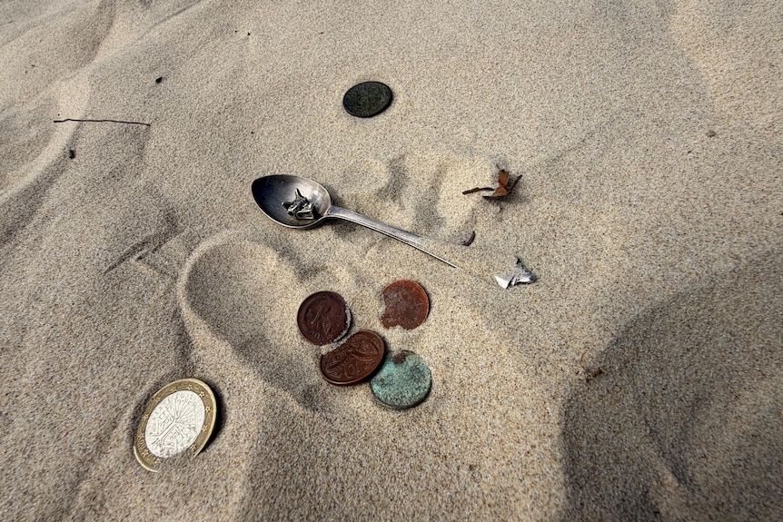 A collection of coins and a teaspoon on the sand at the beach.