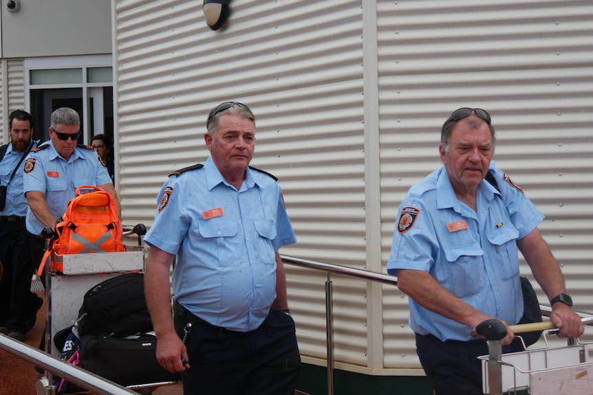 Men in blue uniforms walk down a ramp with baggage in front of a building.