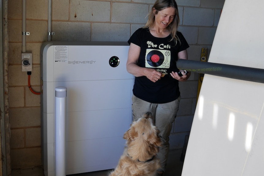 A woman wearing a black t-shirt looks down at a phone in her hand, there's a white battery on the wall behind and a dog in front