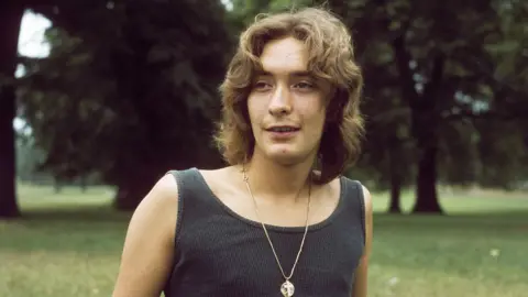 Getty Images Chris Rea pictured in 1979 stood in a park with grass and trees wearing a dark vest and gold neck chain with pendent. He has shoulder length brown hair.