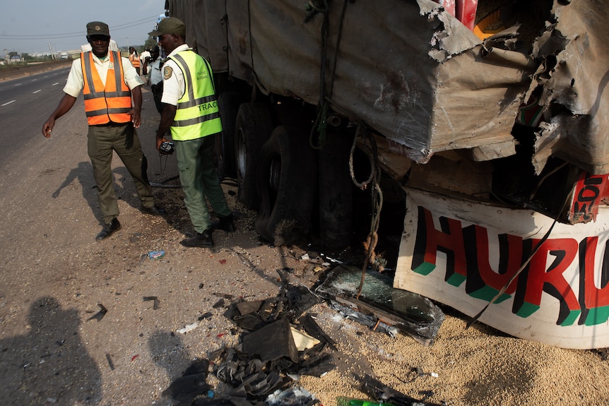 Two Nigerian officials stand next to a truck with the left rear badly damaged after a crash.