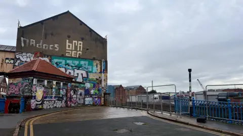 Richard Madden/BBC A street rises towards a riverside. Early 20th century blue railings, lamp posts and a five-sided bridge control building, which is covered in graffiti can be seen. More graffiti is painted on the wall of a large warehouse. On the far bank of the river, a lorry can be seen parked and another old brick warehouse stands in the distance.