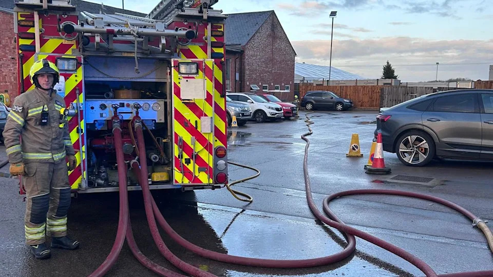 A firefighter stands at the rear of one of the fire engines which has four hoses leading off towards the building.