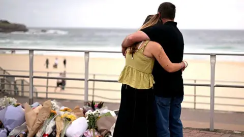 Getty A couple with their backs to the camera embrace in front a floral tribute on the promenade with the beach and ocean in the background