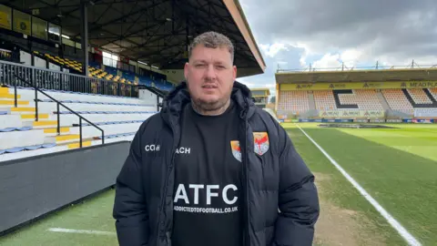 Charlie Barker, a man with blonde curly hair, is wearing a branded black coat and t-shirt featuring the name of his group 'Addicted to Football' while standing on the pitch at an empty football stadium.