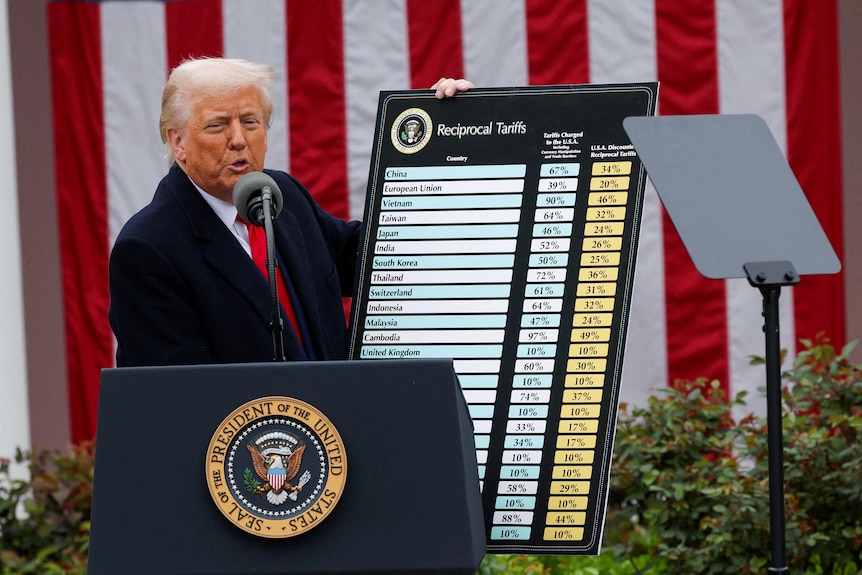 Man in a suit speaking into a microphone behind a podium while holding up a chart