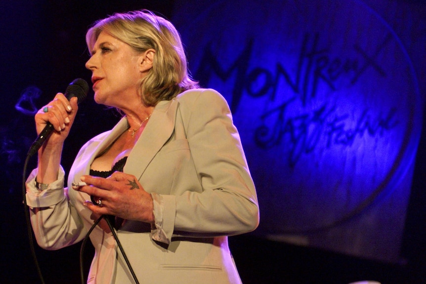 Marianne Faithfull wearing a creme-coloured blazer and singing into a handheld microphone in front of a blue and black backdrop