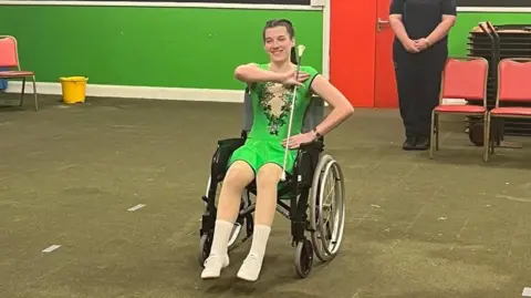 Supplied Hannah wearing a green majorettes outfit and holding a majorette baton while sitting in a wheelchair. 