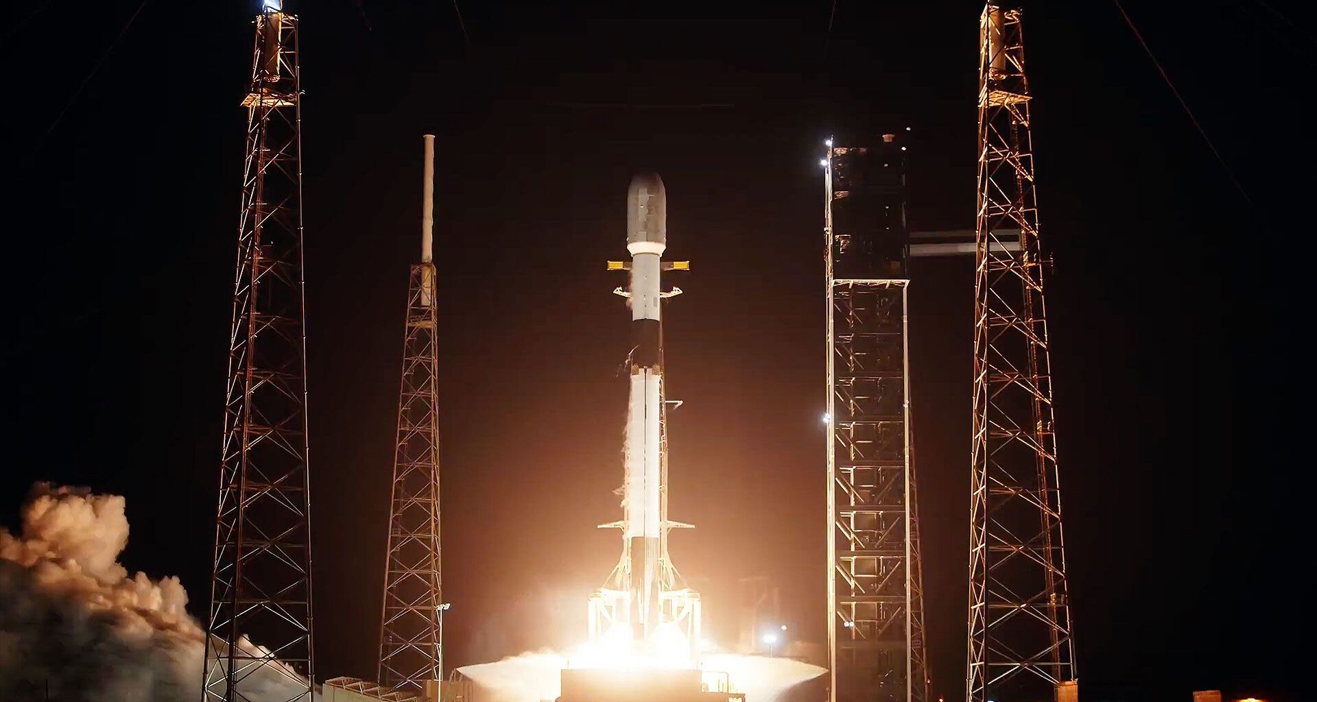 a white and black rocket lifts off into the darkness of night, its bright orange-tinted plume lighting up its launch pad.