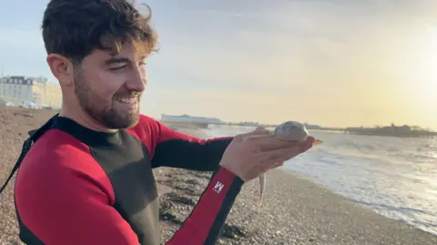 Hannah Roe / BBC Gio, a young man with brown hair in a red and black wetsuit proudly holds a small cat shark up to the camera.