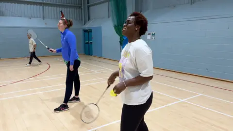 Two women stand on the badminton court playing a doubles match. Sam is closest to the camera with Georgina further away and wearing a headband decorated with Santa's legs sticking out of a chimney.