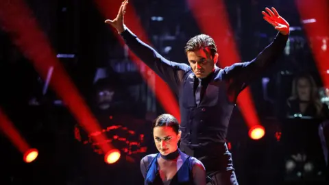 PA Media Amber Davies is crouched in front of Nikita Kuzmin during a Strictly performance. He is wearing a purple waistcoat and tie with her in a matching dress. The lights and audience around the stage can be seen in the background.