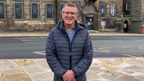 Jon Plant - wearing glasses, and a blue coat - stands and smiles on a town square. Behind him is a road and a large, old building.